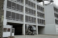A multi-story parking garage with a large truck parked at the entrance. The facade consists of open metal grills and concrete, with safety markings on some pillars. Staircases are visible on the side of the structure, and there is a small security booth near the entrance.