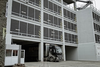 A multi-story parking garage with a large truck parked at the entrance. The facade consists of open metal grills and concrete, with safety markings on some pillars. Staircases are visible on the side of the structure, and there is a small security booth near the entrance.