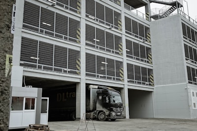 A multi-story parking garage with a large truck parked at the entrance. The facade consists of open metal grills and concrete, with safety markings on some pillars. Staircases are visible on the side of the structure, and there is a small security booth near the entrance.