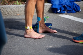 A person stands barefoot on a paved surface, wearing a yellow strap around the ankle and blue athletic tape on the leg. Nearby, a pair of navy blue running shoes with lime green laces is placed, and a pile of clothes in shades of blue lies on the ground.