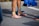 A person stands barefoot on a paved surface, wearing a yellow strap around the ankle and blue athletic tape on the leg. Nearby, a pair of navy blue running shoes with lime green laces is placed, and a pile of clothes in shades of blue lies on the ground.