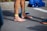 A person stands barefoot on a paved surface, wearing a yellow strap around the ankle and blue athletic tape on the leg. Nearby, a pair of navy blue running shoes with lime green laces is placed, and a pile of clothes in shades of blue lies on the ground.