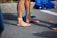 A person stands barefoot on a paved surface, wearing a yellow strap around the ankle and blue athletic tape on the leg. Nearby, a pair of navy blue running shoes with lime green laces is placed, and a pile of clothes in shades of blue lies on the ground.
