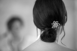 A close-up view of a woman's dark hair styled in a low bun with an ornamental hairpin adorned with pearls and intricate metallic design. The background is softly blurred, creating a depth effect.