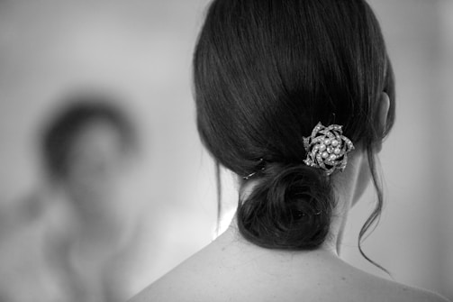 A close-up view of a woman's dark hair styled in a low bun with an ornamental hairpin adorned with pearls and intricate metallic design. The background is softly blurred, creating a depth effect.