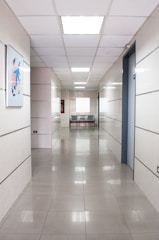 A long, clean corridor with smooth tiled floors and white walls, illuminated by bright ceiling lights. There is a piece of abstract artwork mounted on the left wall and a pair of metallic chairs at the end of the corridor, next to an emergency fire hose cabinet.