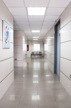 A long, clean corridor with smooth tiled floors and white walls, illuminated by bright ceiling lights. There is a piece of abstract artwork mounted on the left wall and a pair of metallic chairs at the end of the corridor, next to an emergency fire hose cabinet.