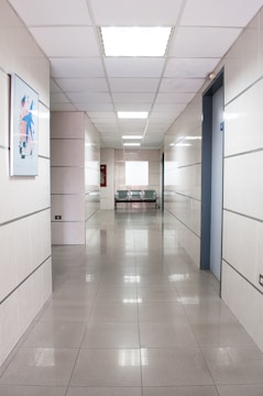 A long, clean corridor with smooth tiled floors and white walls, illuminated by bright ceiling lights. There is a piece of abstract artwork mounted on the left wall and a pair of metallic chairs at the end of the corridor, next to an emergency fire hose cabinet.