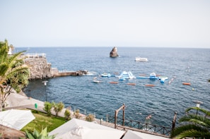 A serene coastal view featuring a vibrant blue sea with a prominent rocky formation emerging from the water. A floating water park with inflatable structures is set up in the ocean, attracting swimmers and visitors. The foreground includes lush greenery, palm trees, and a stone pathway leading down to the water. A boat is visible in the distance, contributing to the picturesque and lively setting.