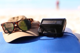 A pair of tortoiseshell sunglasses rests on a beige hat, next to a black electronic device labeled 'Control D Blood Glucose Monitor'. These items are placed on a blue fabric surface, with a blurred background suggesting a sunny beach setting.