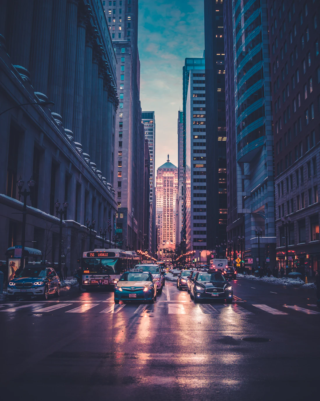 City street at dusk, office towers and headlights reflected on wet asphalt