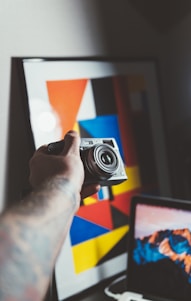 A hand holding a camera is positioned in front of a modern art piece with geometric shapes and vibrant colors. A laptop is partially visible in the foreground, displaying a mountain landscape on the screen.