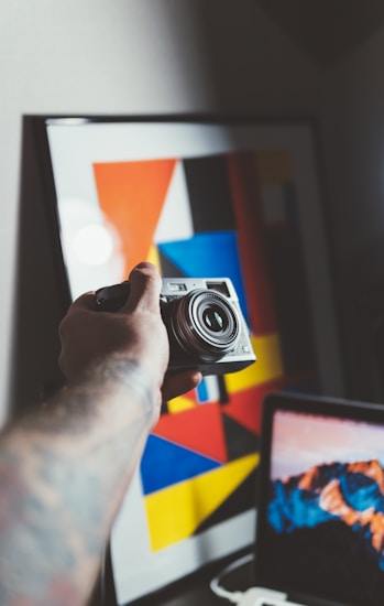 A hand holding a camera is positioned in front of a modern art piece with geometric shapes and vibrant colors. A laptop is partially visible in the foreground, displaying a mountain landscape on the screen.