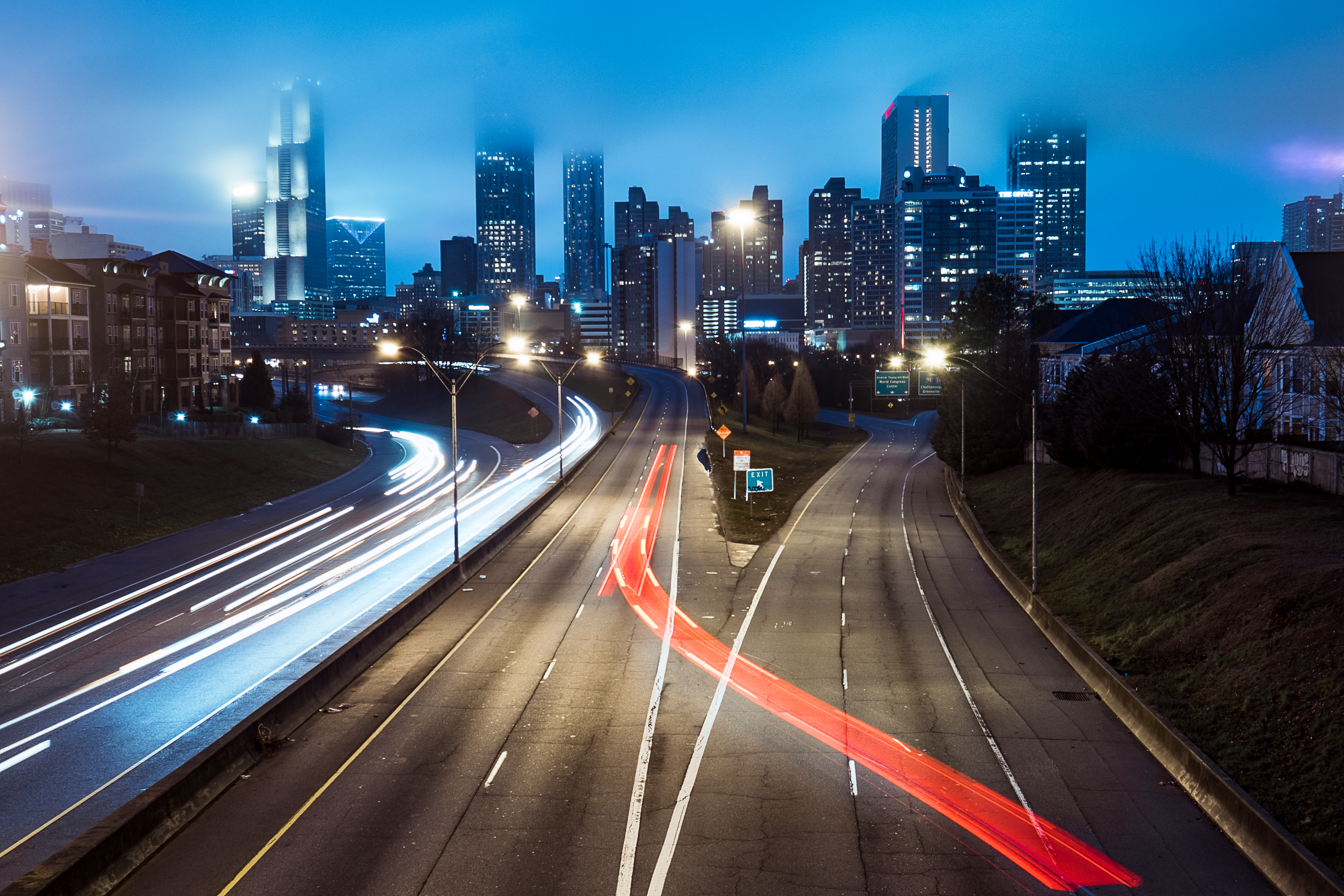 Am empty highway leading to a light downtown at night.  There are blue lights on the left and on the right is a divided lane with a red light cutting over avoiding the exit.