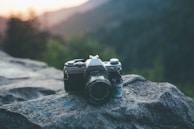 A vintage camera rests on a large rock, with a roll of film positioned beside it. The background features a soft-focus view of mountains and trees with the warm glow of sunset.