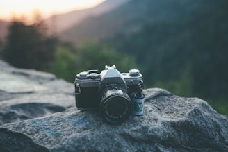 A vintage camera rests on a large rock, with a roll of film positioned beside it. The background features a soft-focus view of mountains and trees with the warm glow of sunset.