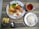 A meal is arranged on a table with a main plate featuring a variety of foods including salmon, a sausage, mixed vegetables, a fried egg, and potatoes. A smaller serving of plain white rice is placed on a separate dish. A cup contains a light-colored tea, and there are slices of apple in a glass bowl. Cutlery is neatly positioned next to the main plate, all on a striped tablecloth.