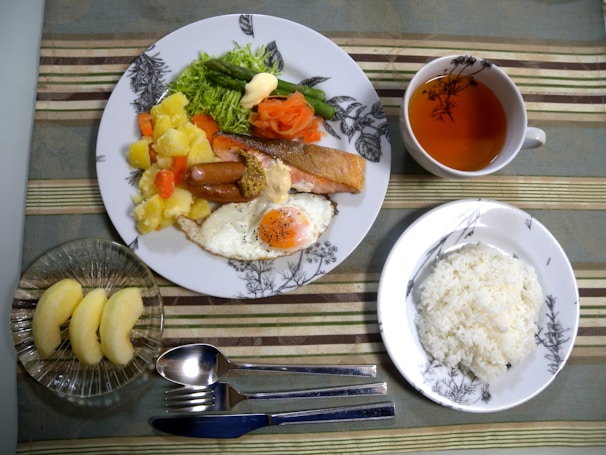 A meal is arranged on a table with a main plate featuring a variety of foods including salmon, a sausage, mixed vegetables, a fried egg, and potatoes. A smaller serving of plain white rice is placed on a separate dish. A cup contains a light-colored tea, and there are slices of apple in a glass bowl. Cutlery is neatly positioned next to the main plate, all on a striped tablecloth.