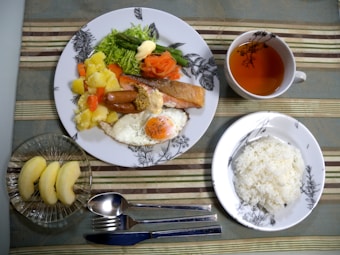 A meal is arranged on a table with a main plate featuring a variety of foods including salmon, a sausage, mixed vegetables, a fried egg, and potatoes. A smaller serving of plain white rice is placed on a separate dish. A cup contains a light-colored tea, and there are slices of apple in a glass bowl. Cutlery is neatly positioned next to the main plate, all on a striped tablecloth.