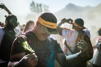 A person covered in colored powder is participating in a color run event. The individual is wearing a yellow visor with 'COLOR RUNNER' written on it and appears joyful and energetic. The background shows other participants, some taking photos, all amidst a cloud of multicolored dust. Sunlight filters through the scene, adding a vivid and dynamic atmosphere.