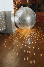 A large disco ball rests on a wooden floor, reflecting small squares of light in a pattern across the room. The light creates a dynamic, lively atmosphere. A wine bottle is visible in the background, along with a bicycle wheel and other household objects, suggesting a casual, lived-in space.