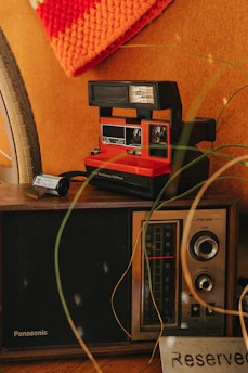 A vintage setup featuring a retro Polaroid camera, an old-fashioned radio with a tuning dial, and a reserved sign. A bicycle wheel and a crocheted orange fabric are partially visible, adding to the nostalgic ambiance.