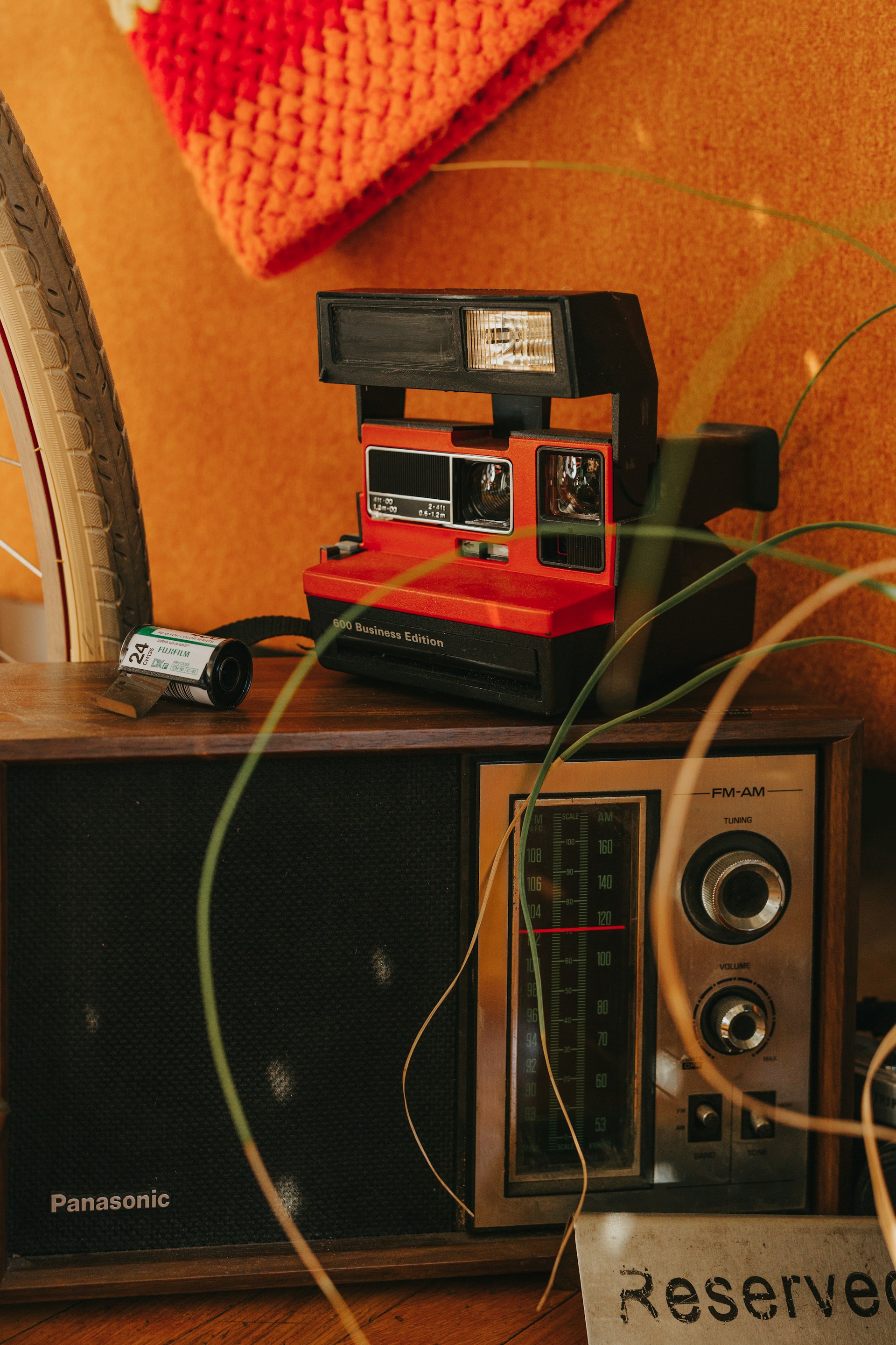 A vintage setup featuring a retro Polaroid camera, an old-fashioned radio with a tuning dial, and a reserved sign. A bicycle wheel and a crocheted orange fabric are partially visible, adding to the nostalgic ambiance.