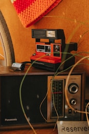 A vintage setup featuring a retro Polaroid camera, an old-fashioned radio with a tuning dial, and a reserved sign. A bicycle wheel and a crocheted orange fabric are partially visible, adding to the nostalgic ambiance.