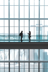 Two silhouetted figures stand facing each other on a walkway inside a building with large floor-to-ceiling windows. The spacious environment is filled with natural light, creating a serene atmosphere. The background reveals a snowy outdoor scene visible through the windows.