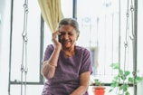 An elderly woman with gray hair is smiling joyfully while talking on a mobile phone. She is seated indoors with light flowing in through a window behind her, partially covered by a curtain. The background includes a metal grill, some green plants, and a small red pot.