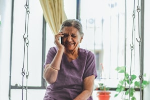 An elderly woman with gray hair is smiling joyfully while talking on a mobile phone. She is seated indoors with light flowing in through a window behind her, partially covered by a curtain. The background includes a metal grill, some green plants, and a small red pot.