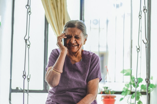 An elderly woman with gray hair is smiling joyfully while talking on a mobile phone. She is seated indoors with light flowing in through a window behind her, partially covered by a curtain. The background includes a metal grill, some green plants, and a small red pot.