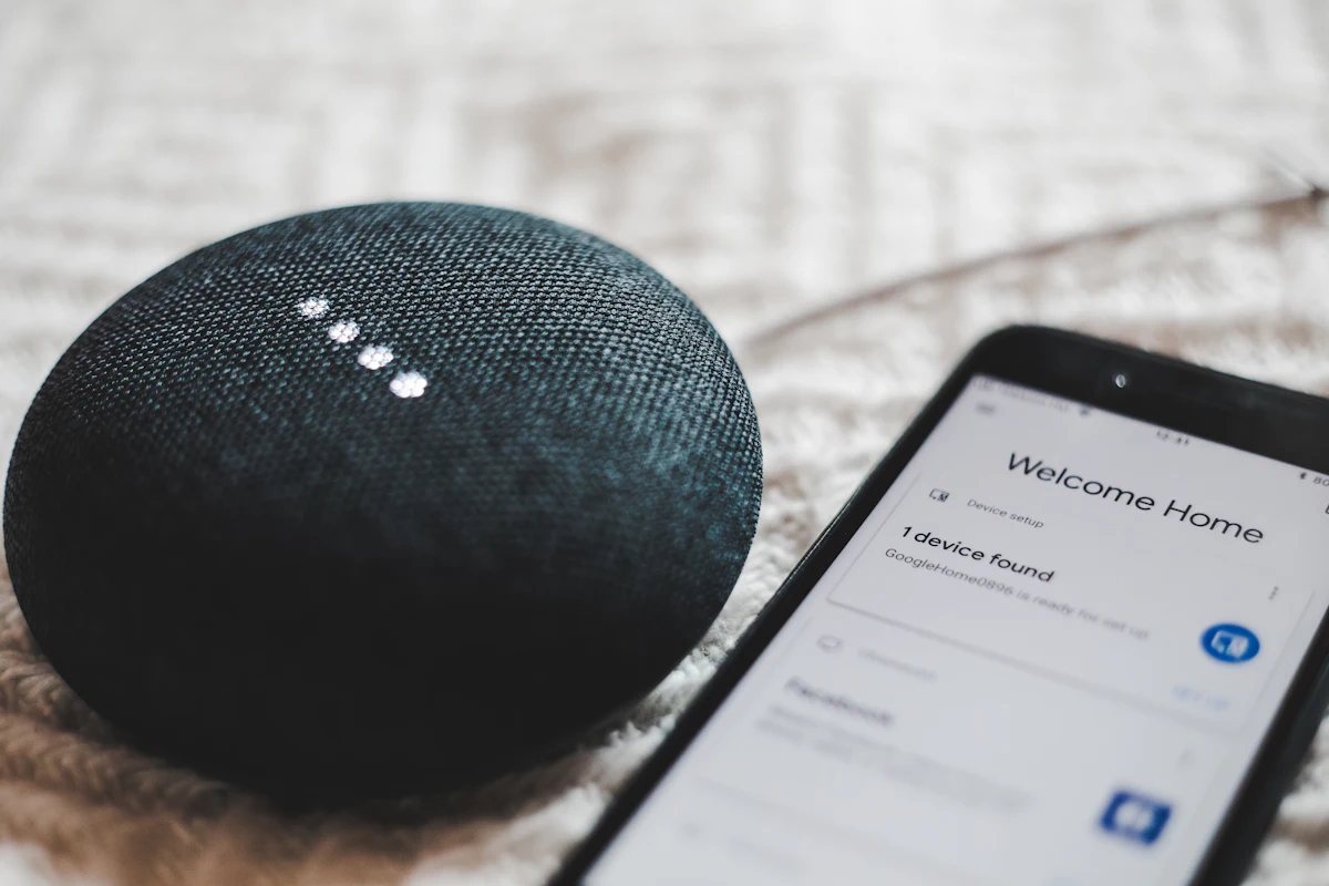 Google Home Mini berwarna abu-abu di samping smartphone sebagai pusat kontrol perangkat smart home rumahan