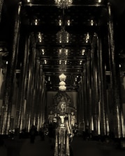 A richly decorated interior of a temple featuring tall, ornately carved pillars and multiple chandeliers hanging from the ceiling. In the center stands a large golden statue of a deity, with additional statues visible in the background. The space is dimly lit, creating a serene and solemn atmosphere.