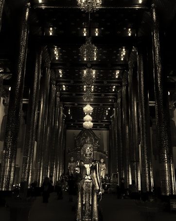 A richly decorated interior of a temple featuring tall, ornately carved pillars and multiple chandeliers hanging from the ceiling. In the center stands a large golden statue of a deity, with additional statues visible in the background. The space is dimly lit, creating a serene and solemn atmosphere.