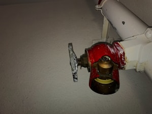 A red valve is attached to a white pipe on a textured wall. The valve appears metallic with visible wear, indicating regular use or age. The valve handle is tarnished and somewhat rusty, contrasting with the bright red hue of the main valve body.