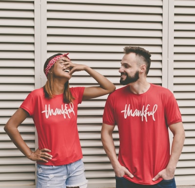 A man and woman are standing side by side in front of a slatted wall, both wearing red t-shirts with the word 'thankful' printed in white. The woman is wearing a red headband and denim shorts, and she appears to be laughing and covering her eyes with her hand. The man has a beard and is smiling as he looks at her.