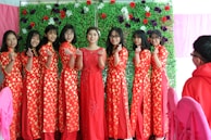 A group of women stand in a line wearing traditional red dresses with yellow polka dots. They are smiling and making a hand gesture with their fingers forming hearts. The background consists of a vibrant green wall adorned with colorful flowers. Pink chairs are visible in the foreground, implying a festive setting.