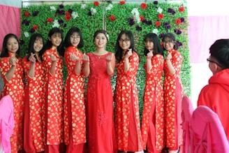 A group of women stand in a line wearing traditional red dresses with yellow polka dots. They are smiling and making a hand gesture with their fingers forming hearts. The background consists of a vibrant green wall adorned with colorful flowers. Pink chairs are visible in the foreground, implying a festive setting.