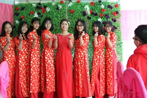A group of women stand in a line wearing traditional red dresses with yellow polka dots. They are smiling and making a hand gesture with their fingers forming hearts. The background consists of a vibrant green wall adorned with colorful flowers. Pink chairs are visible in the foreground, implying a festive setting.