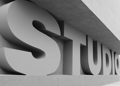 Large, three-dimensional letters spelling out the word 'STUDIO' are positioned against a modern architectural structure. The letters are bold and cast shadows due to a light source from the side, and the overall color scheme is monochrome.