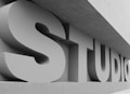 Large, three-dimensional letters spelling out the word 'STUDIO' are positioned against a modern architectural structure. The letters are bold and cast shadows due to a light source from the side, and the overall color scheme is monochrome.
