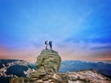 Two hikers stand atop a rocky cliff, surrounded by expansive mountainous terrain. The sky is clear with a gradient from blue to warmer hues near the horizon. The scene conveys a sense of adventure and exploration.