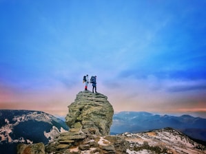 Two hikers stand atop a rocky cliff, surrounded by expansive mountainous terrain. The sky is clear with a gradient from blue to warmer hues near the horizon. The scene conveys a sense of adventure and exploration.