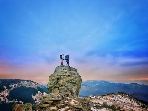 Two hikers stand atop a rocky cliff, surrounded by expansive mountainous terrain. The sky is clear with a gradient from blue to warmer hues near the horizon. The scene conveys a sense of adventure and exploration.
