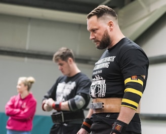 Three people are in a gym-like setting. Two men in the foreground are wearing athletic gear, including belts and wristbands, with focused expressions. A woman in the background is casually dressed in a pink sweatshirt, standing with arms crossed.