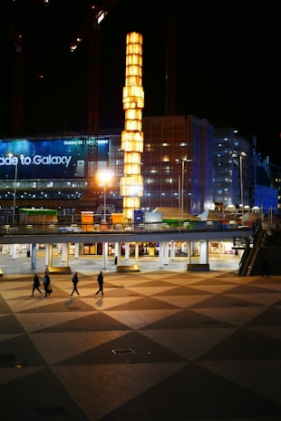 A brightly lit vertical structure towers over an urban setting, surrounded by construction cranes and a large building with illuminated signage advertising a smartphone. In the foreground, a few pedestrians walk across a patterned plaza.