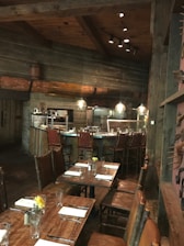 A rustic dining area with wooden tables and chairs, set up for service. The tables are adorned with glassware, silverware, folded napkins, and small floral arrangements in jars. The setting features wooden walls and ceiling beams, with low-hanging pendant lights providing warm illumination. In the background, there is a bar area with high-backed woven bar stools.