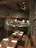 A rustic dining area with wooden tables and chairs, set up for service. The tables are adorned with glassware, silverware, folded napkins, and small floral arrangements in jars. The setting features wooden walls and ceiling beams, with low-hanging pendant lights providing warm illumination. In the background, there is a bar area with high-backed woven bar stools.