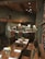 A rustic dining area with wooden tables and chairs, set up for service. The tables are adorned with glassware, silverware, folded napkins, and small floral arrangements in jars. The setting features wooden walls and ceiling beams, with low-hanging pendant lights providing warm illumination. In the background, there is a bar area with high-backed woven bar stools.